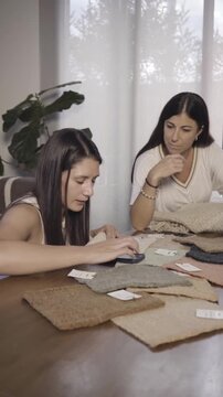 Two women lean over a table scattered with woven fabric samples, discussing texture and pattern in soft natural light. Their