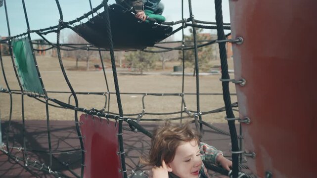 Toddler boy climbing and sitting on modern rope playground structure at suburban park