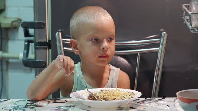 A thoughtful little boy pauses while eating a plate of spaghetti. He sits quietly at a kitchen table, looking curious and content during his mealtime.