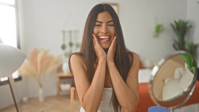 Woman cups cheeks with hands at vanity mirror in a bright studio, smiling with playful expression and visible makeup on face; self care joy.