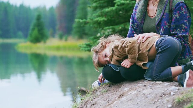 Toddler boy cuddling on mother's lap beside calm mountain lake with pine forest