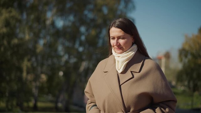 Close up of woman walking outdoors in urban park with trees and buildings visible. Woman wears brown coat tied at waist cream scarf long dark hair carrying handbag hands partially tucked