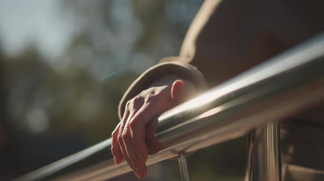 Close up of woman hand resting on metal railing with sunlight reflection. subject wears brown coat showing detailed fabric texture and sleeve cuff. Background is blurred outdoor scene