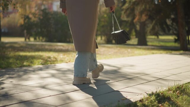 Close up of woman walking outdoors on paved path holding black handbag. Woman wears long brown coat light blue jeans and beige boots with visible shadows from trees in park setting