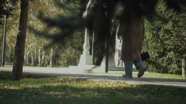 Woman in long coat walks along park path holding small handbag. scene features trees and sunlight filtering through foliage on autumn day with fallen leaves scattered across ground
