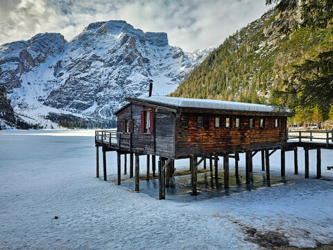 Lake Braies (Pragser See) in South Tyrol in winter, view of snowy mountain and Lake, Snow-covered wooden hut on stilts beside frozen lake. Mountains rise behind, partially snow-capped. 