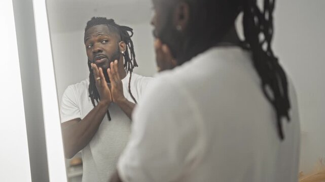 Man adjusting dreadlocks and touching face in lighted mirror in studio while checking skin and hair; self care confidence.