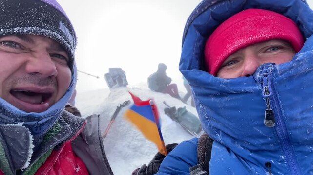 Happy mountaineers posing in close-up on Mount Ararat summit with Armenian flag in stormy snow, showing summit achievement, expedition success, extreme weather endurance, and adventure travel