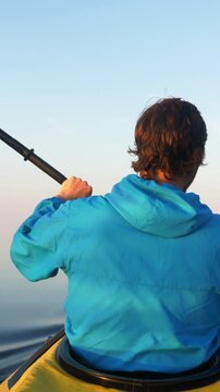 Young man in a blue hooded jacket kayaking on a tranquil lake at dawn, with the warm sunlight reflecting on his hair as he paddles across the smooth water in a yellow boat