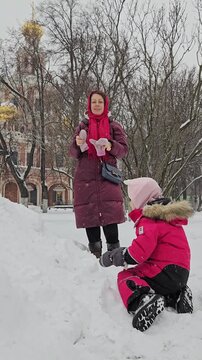 Woman and child enjoy snowy park together. They build snow pile with hands. Child sits on snow, watches woman. Snowy trees stand behind them. Winter light shines on their faces