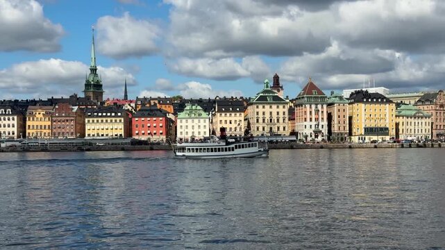Vintage passenger boat cruising across the harbor in front of the colorful historic architecture and church spire of Gamla Stan in Stockholm, Sweden.