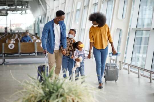 A Black family with two children wearing protective face masks and carrying suitcases walks through a modern airport terminal