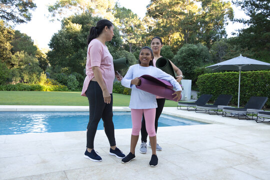 Diverse female family with youth standing holding rolled yoga mats in athletic wear on pool deck