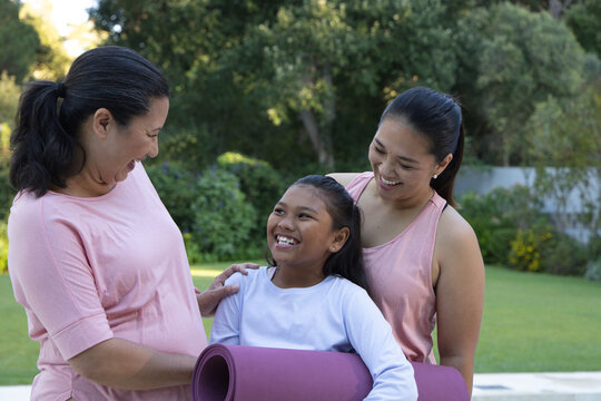 Diverse female family smiling laughing in backyard in pink and white tops holding purple yoga mat