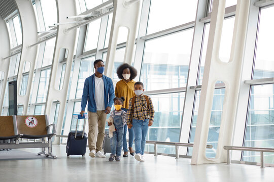 A diverse family of four wearing protective face coverings walks together through a modern airport terminal with their travel bags