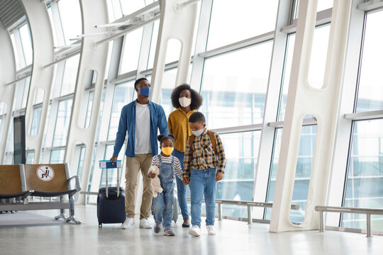 A diverse family with children walks together through a modern airport terminal, all wearing protective face masks for travel safety