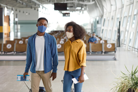 A man and woman wearing protective masks walk through a modern airport terminal, holding travel documents and luggage
