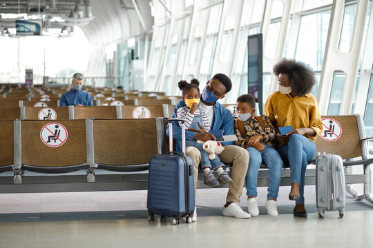 A diverse family with children sits in an airport terminal, all wearing face masks and observing social distancing protocols while waiting for their flight