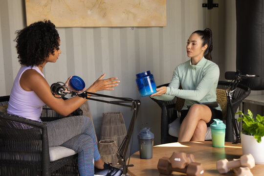 Two women in workout gear sitting in studio exchanging blue supplement tub recording with boom mics