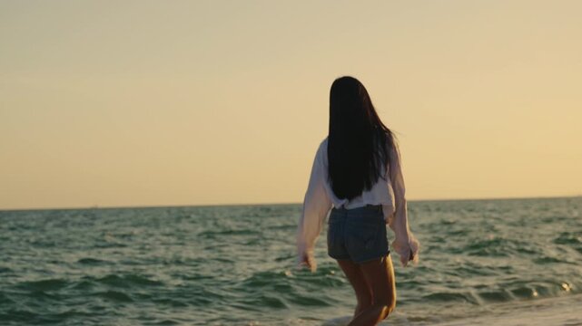 Happy young woman enjoying summer walk on sea sand beach at sunset, serene ocean sunrise travel lifestyle concept showing carefree female in casual clothes during vacation trip.