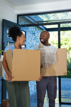 Couple carrying cardboard moving boxes in home entryway, smiling and holding fringed throw