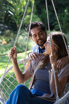 Diverse couple sitting on macrame hanging swing in backyard, both wearing jeans and holding ropes