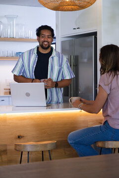 Diverse couple chatting across wood-front kitchen island at home holding mugs and laptop