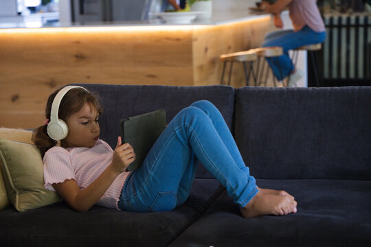 Mother and child reclining on dark sofa at home, child wearing pink top headphones holding tablet