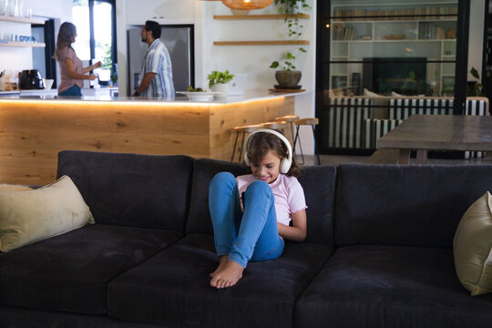 Family girl sitting on sofa at home wearing headphones using phone while parents talking at island