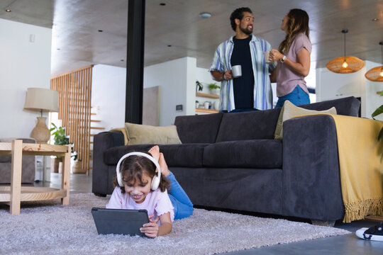 African American family talking by sofa while child lying on gray rug wearing headset using tablet