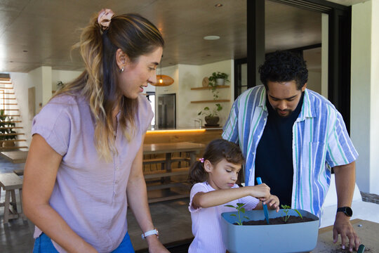 Diverse family watching youth girl planting seedlings in planter with blue trowel on table at home