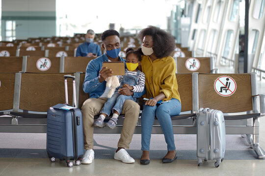 A family with a young child sits together in an airport waiting area, all wearing protective face masks and looking at a tablet device