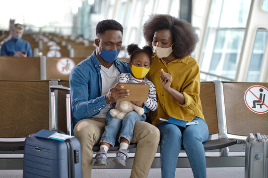 A father, mother, and daughter sit together in an airport terminal, all wearing protective face coverings and looking at a digital device