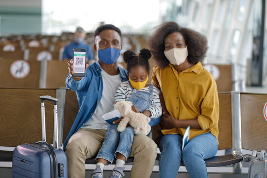 A Black family wearing face masks sits in an airport waiting area with luggage, displaying a digital health pass on a smartphone for travel