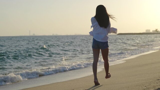 Happy beautiful woman walking on wet sand beach enjoying sunset day of leisure and freedom, strolling along coastline as gentle ocean waves wash ashore during warm summer evening vacation.
