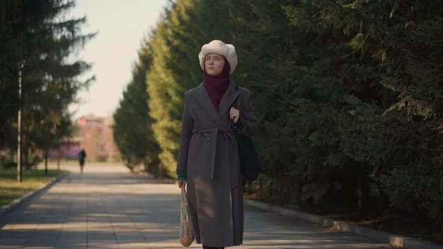 Woman walks along tree lined path carrying bags dressed in warm clothing. Woman holds items including mesh produce bags and wears boots with visible texture detail. scene features outdoor