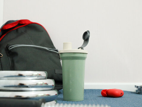 Fitness Gear Arranged On Carpet Beside Black Backpack, Green Shaker Bottle, Chrome Dumbbells, And Red Earbuds, Creating Clean Indoor Workout Still Life With Copy Space For Exercise Planning