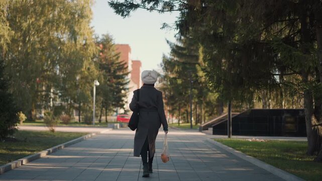 Woman walks steadily down tree lined path in urban park carrying bags and wearing winter clothing. scene features soft natural light filtering through tall trees with green foliage on both