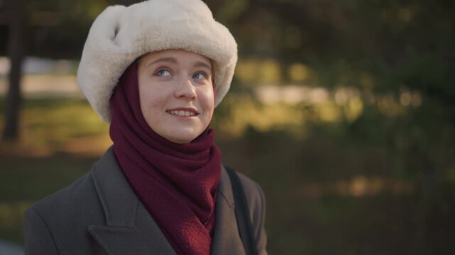 Close up of woman wearing traditional winter clothing including white fur hat and maroon scarf standing outdoors in soft natural light. Her attire suggests cultural or regional significance