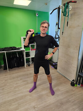 Man holds kettlebell in fitness studio. He wears black athletic gear and purple socks. Green wall and wooden floor are visible. Equipment hangs on the wall behind him electro muscle stimulation