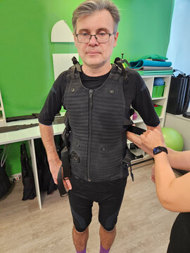 Man wears black vest while standing in fitness studio. Another person adjusts the vest. Room has green walls and exercise equipment. Man wears glasses and black shorts electro muscle stimulation