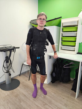 Man wears black resistance suit and purple socks. He stands in fitness studio with walls. Equipment and storage units are visible behind him. He looks directly with glasses electro muscle stimulation