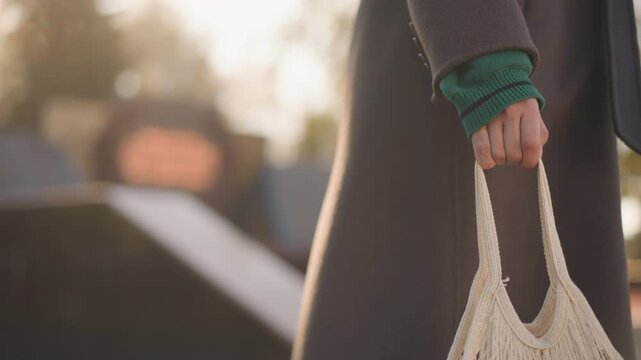Close up of woman walking outdoors holding off white net bag in her right hand and carrying black shoulder bag. scene features soft natural light with warm tones focusing on lower half