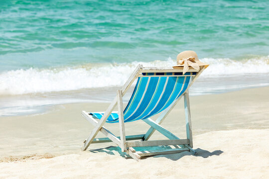 Beach chair with blue and white stripes and a straw hat resting on it, positioned on sandy shore near gentle ocean waves under bright sunlight. Summer tropical vacation and relax concept. Copy space