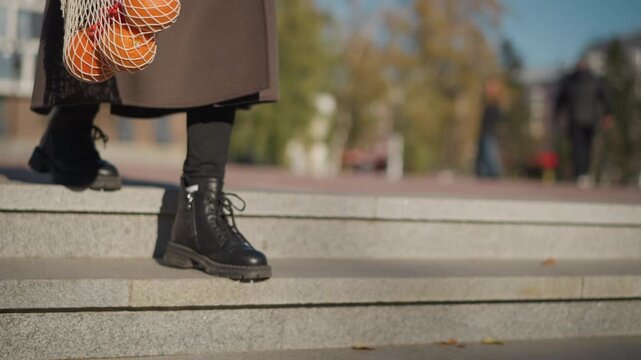 Close up of woman walking down stone steps outdoors carrying net bags with oranges and red berries. Woman wears black boots under long brown coat transitioning from paved ground to higher