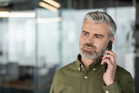 Serious grey-haired businessman listening intently on a smartphone during a phone call, standing in a contemporary office setting with blurred background