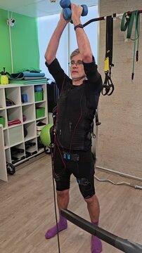 Man exercises with resistance garment in gym setting. He lifts dumbbells overhead while wearing the gear. His posture shows focused movement during workout session electro muscle stimulation
