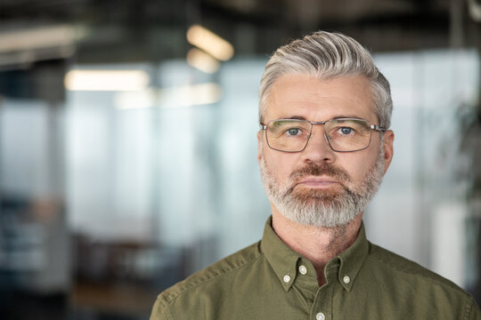 Serious mature businessman with grey hair and beard, wearing glasses and green shirt, standing waist-up in a modern office, direct gaze conveying confidence and thoughtful leadership