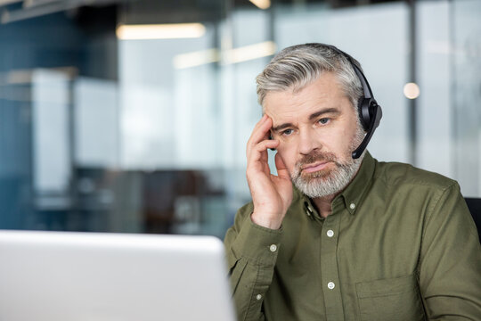 Stressed mature businessman wearing a headset and touching his head while sitting at a desk with a laptop, looking tired and overwhelmed in a contemporary office environment