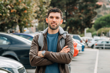 Obraz premium Confident young man with crossed arms standing outdoors in a parking lot on a sunny autumn day, smiling with a casual travel and tourism lifestyle concept